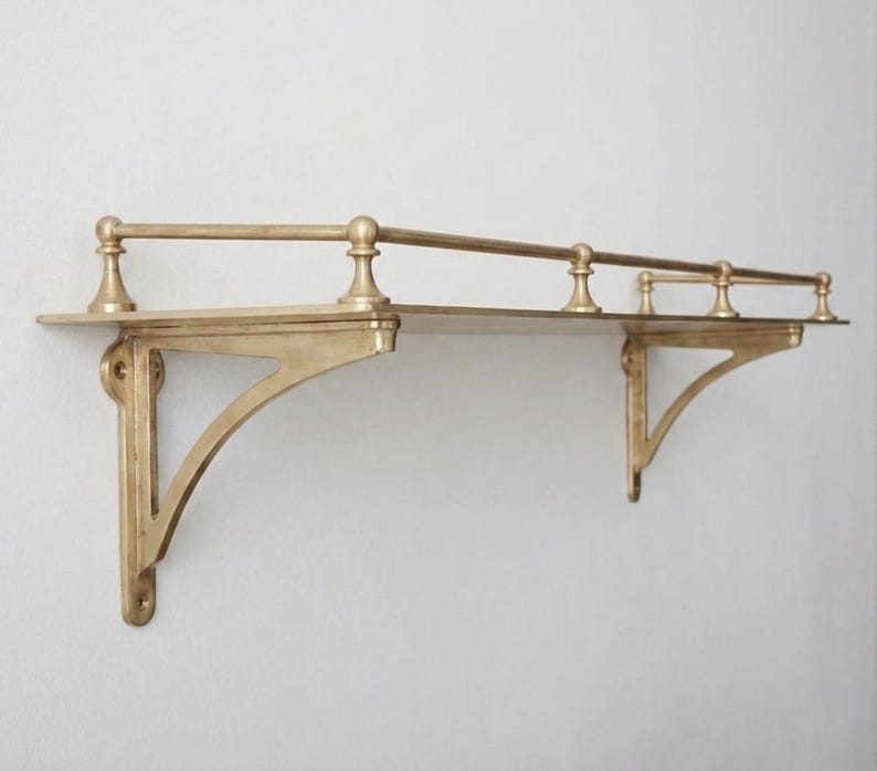 May include: A wall-mounted shelf crafted from brass with a metallic gold finish. The shelf features a flat rectangular surface supported by two decorative triangular brackets and includes a protective railing along the back and sides with four posts.