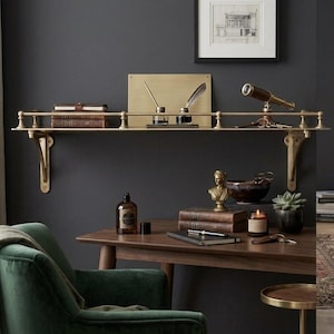 May include: A wall-mounted brass shelf with a railing, supported by two ornate brackets. The shelf holds two stacked books, a rectangular brass plate, two inkwells with quill pens, and a small brass telescope with a set of keys.