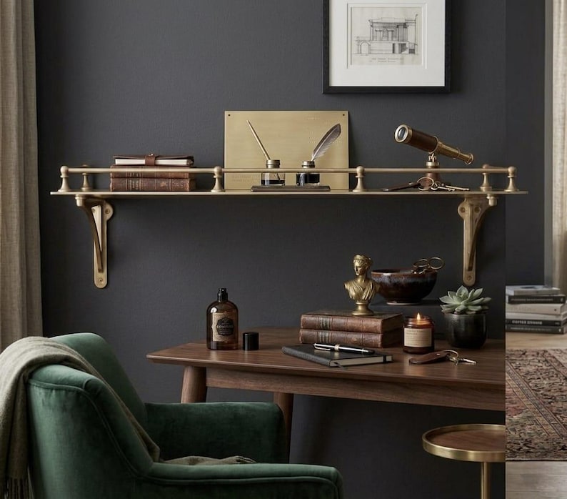 May include: A wall-mounted brass shelf with a railing, supported by two ornate brackets. The shelf holds two stacked books, a rectangular brass plate, two inkwells with quill pens, and a small brass telescope with a set of keys.