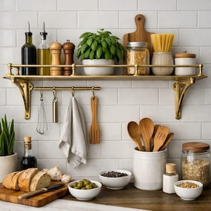 May include: A brass kitchen shelf with a rail holds two glass oil bottles, two wooden salt and pepper grinders, a potted green plant, a glass jar, a ceramic container with pasta, and a white canister. A hanging rack displays a whisk and towel.