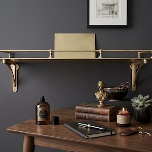 May include: A wall-mounted brass shelf featuring a horizontal railing supported by two decorative brackets. The shelf has a flat surface and a rectangular brass backplate. The metal has a brushed finish, providing a metallic gold appearance.