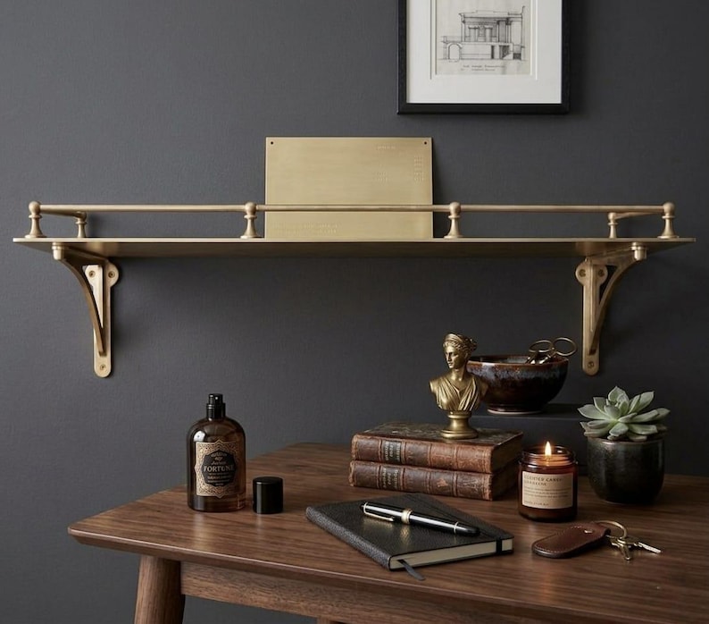 May include: A wall-mounted brass shelf featuring a horizontal railing supported by two decorative brackets. The shelf has a flat surface and a rectangular brass backplate. The metal has a brushed finish, providing a metallic gold appearance.