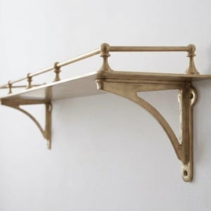 May include: A wall-mounted shelf made of brass with a metallic gold finish. The shelf features two decorative support brackets and a horizontal railing along the front and sides, held in place by four small vertical posts attached to the flat surface.