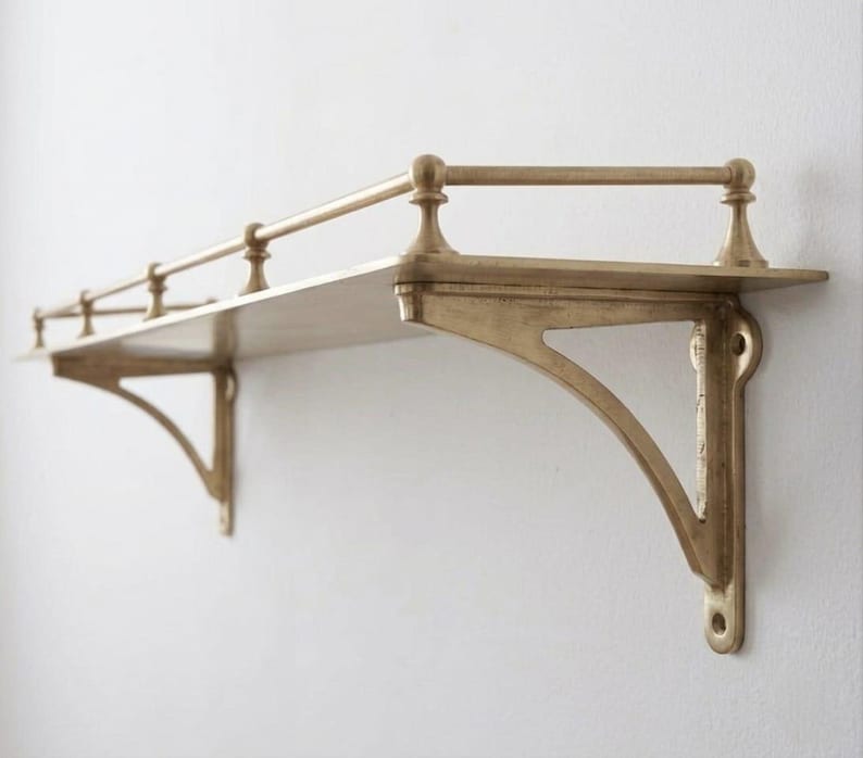 May include: A wall-mounted shelf made of brass with a metallic gold finish. The shelf features two decorative support brackets and a horizontal railing along the front and sides, held in place by four small vertical posts attached to the flat surface.