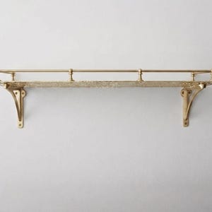 May include: A wall-mounted gold-toned metal shelf featuring a textured, patterned surface and a thin horizontal railing supported by three small vertical posts. The shelf is held in place by two decorative, curved brackets on either side.