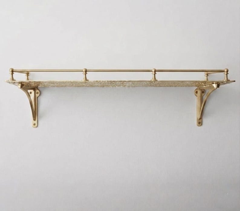 May include: A wall-mounted gold-toned metal shelf featuring a textured, patterned surface and a thin horizontal railing supported by three small vertical posts. The shelf is held in place by two decorative, curved brackets on either side.