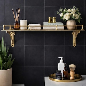 May include: A gold-toned metal wall shelf with a railing and decorative brackets. The shelf holds a reed diffuser, a white candle, a folded white cloth, two gold binoculars, two books, and a vase of white roses. Below, a gold tray holds toiletries.