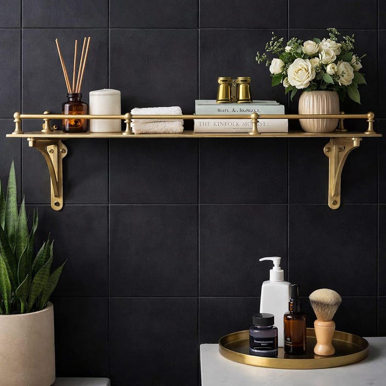 May include: A gold-toned metal wall shelf with a railing and decorative brackets. The shelf holds a reed diffuser, a white candle, a folded white cloth, two gold binoculars, two books, and a vase of white roses. Below, a gold tray holds toiletries.