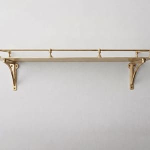 May include: A wall-mounted shelf made of polished brass with a flat rectangular surface. The shelf features two decorative support brackets on the ends and a thin horizontal railing along the back edge, supported by three small vertical posts.