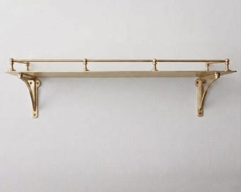 May include: A wall-mounted shelf made of polished brass with a flat rectangular surface. The shelf features two decorative support brackets on the ends and a thin horizontal railing along the back edge, supported by three small vertical posts.
