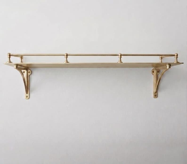 May include: A wall-mounted shelf made of polished brass with a flat rectangular surface. The shelf features two decorative support brackets on the ends and a thin horizontal railing along the back edge, supported by three small vertical posts.