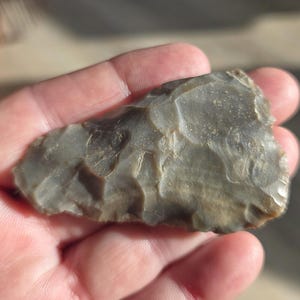 May include: A grey, chipped stone tool, possibly a prehistoric arrowhead or scraper, held in a hand. The stone has a rough, textured surface with various shades of grey and brown. The tool's edges are jagged, indicating it was likely shaped by hand.
