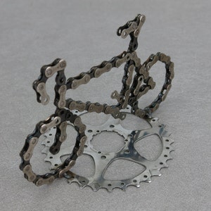 May include: A bicycle sculpture made entirely of silver bike chains. The sculpture is of a bicycle with two wheels, handlebars, and a seat. The sculpture is on a silver metal base.