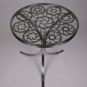 Puede incluir: Una mesa redonda de vidrio con una base cromada. La superficie de la mesa está decorada con un patrón de piñones de bicicleta.