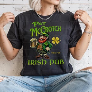 Op de afbeelding: Zwart t-shirt met een groene en gele afbeelding. De afbeelding toont een cartoon kabouter met een biertje, de tekst "Pat McCrotch Irish Pub" en een klavertjevier. De tekst "Since '69" is ook opgenomen.