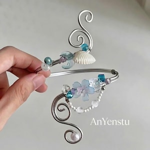 May include: A silver-toned arm cuff with decorative swirls and floral accents. The cuff features blue and white flowers, pearls, and a seashell. The text "AnYenstu" is visible on the cuff.