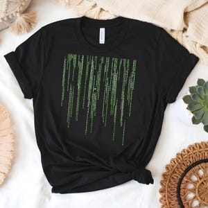 May include: Black t-shirt featuring a green digital rain design. The graphic consists of vertical lines of green characters, resembling code. The shirt is made of a soft material and has a classic crew neck.