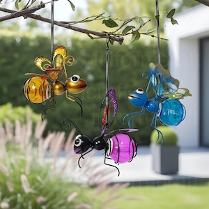 May include: Three hanging metal insect decorations. One is gold and orange, another is black and pink, and the third is blue and green. The insects are hanging from a tree branch with a blurred green background.