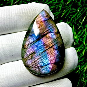 May include: A teardrop-shaped labradorite gemstone with a polished surface. The stone displays a vibrant play of color, including blues, purples, and oranges, with dark linear inclusions. The gemstone is held against a white fabric background.