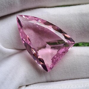 May include: A faceted, triangular-shaped pink gemstone with a transparent, light-reflecting surface. The stone features sharp, geometric edges and a polished finish, showcasing a clear, vibrant pink hue throughout its crystalline structure.