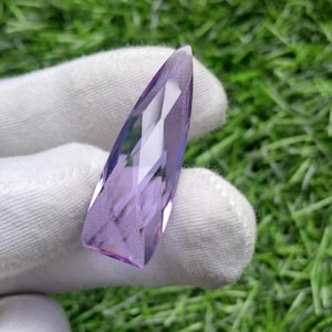 May include: A faceted, elongated, teardrop-shaped amethyst gemstone with a translucent, light purple hue. The crystal features multiple geometric cut surfaces that reflect light, creating a sharp, pointed tip and a wider, flat base.