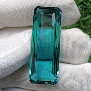 Pode incluir: Uma pedra preciosa retangular com corte esmeralda, de cor azul-esverdeada vibrante e alta clareza. A pedra apresenta bordas facetadas precisas que refletem a luz, exibindo um tom profundo e saturado em toda a sua estrutura alongada.