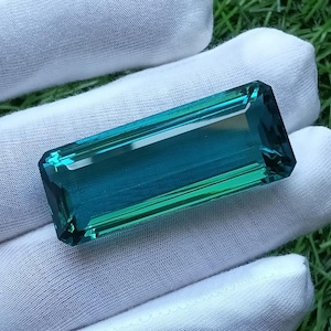 Pode incluir: Uma pedra preciosa retangular facetada com um gradiente vibrante de cores azul e verde-azulado. A pedra apresenta bordas afiadas e limpas e uma superfície polida e reflexiva, exibindo uma saturação profunda que transita do verde-azulado claro para o azul escuro.