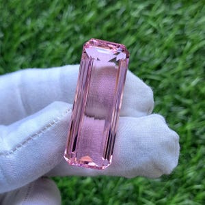 Puede incluir: Una piedra preciosa rosa de corte rectangular con facetas y superficie transparente. La piedra presenta bordes afilados y definidos, con un tono rosa claro y diáfano que muestra un alto nivel de claridad y un acabado pulido que refleja la luz.