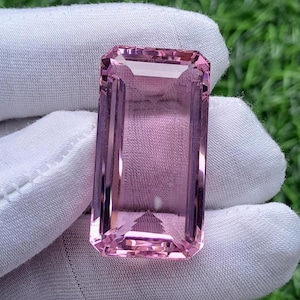 May include: A rectangular, emerald-cut pink gemstone with a transparent, faceted surface. The stone displays a light pink hue and is held against a white fabric background, showcasing its clear, polished edges and geometric symmetry.