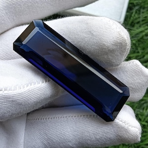 Peut inclure: Une pierre précieuse rectangulaire d'un bleu profond avec une surface à facettes et des bords biseautés nets. La pierre présente une teinte bleu marine translucide avec des surfaces polies qui reflètent la lumière, révélant une coupe géométrique allongée.