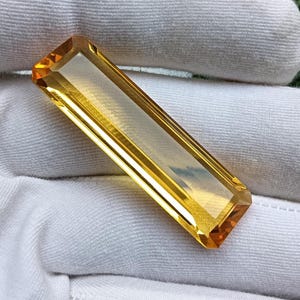 May include: A rectangular, baguette-cut citrine gemstone with a vibrant golden-yellow hue and faceted edges. The stone displays high clarity and a polished surface, reflecting light across its elongated, symmetrical shape and sharp, clean corners.