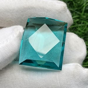May include: A square-cut, translucent teal gemstone with a faceted surface. The stone features a distinct geometric pattern with a central diamond-shaped reflection, showcasing a vibrant blue-green hue and sharp, clean edges throughout the piece.