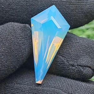 May include: A kite-shaped gemstone with a smooth, faceted surface displaying a translucent light blue color. The stone features sharp, geometric edges and internal light reflections that create a bright, glowing appearance.