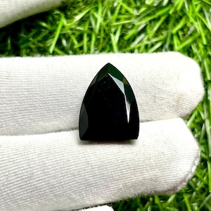 May include: A triangular-shaped, black gemstone with multiple facets. The gem is displayed on a white surface, with a green grassy background. The polished surface of the gem reflects light, highlighting its geometric design.