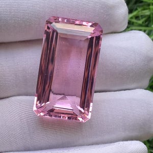 May include: A rectangular, emerald-cut pink gemstone with faceted edges, displaying a translucent and clear appearance. The stone features a light pink hue with subtle variations in colour depth across its surface, reflecting light through its geometric facets.