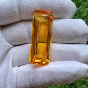 May include: A rectangular, step-cut yellow gemstone with a vibrant, saturated golden-orange hue. The stone features sharp, clean facets that reflect light, creating a brilliant, transparent appearance held against a white fabric background.