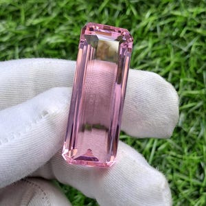 May include: A rectangular, faceted pink gemstone with a clear, transparent appearance and sharp, defined edges. The stone features a step-cut design with multiple facets that reflect light, showcasing a consistent light pink hue throughout the entire piece.