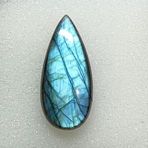 Pode incluir: Uma pedra preciosa de labradorita polida em forma de gota. A pedra exibe uma iridescência azul e verde-azulado vibrante, com fraturas internas criando um padrão único. A pedra preciosa está sobre um fundo branco.