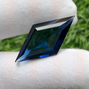 May include: A faceted gemstone with a distinct kite or diamond shape, displaying deep blue and teal hues. The stone features sharp, geometric edges and a polished surface that reflects light, showcasing a clear, transparent quality throughout.