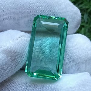 以下が含まれることがあります： 鮮やかで半透明のミントグリーンの色合いを持つ、長方形のエメラルドカットの宝石。石には光を捉えるファセット加工されたエッジがあり、内部の深みが見える透明で反射性の高い表面と、磨き上げられたガラスのような外観が特徴です。