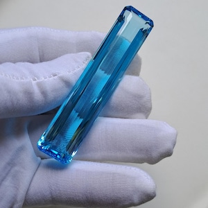 Könnte beinhalten: Ein langer, rechteckiger, facettierter Edelstein in einer leuchtenden, durchscheinenden himmelblauen Farbe. Der Stein weist scharfe, klare Kanten und eine polierte Oberfläche auf, die das Licht reflektiert und seine kristalline Struktur zeigt.