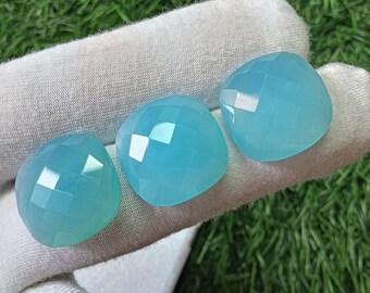 May include: Three faceted, cushion-cut light blue gemstones with a smooth, polished surface. Each stone features a symmetrical geometric pattern of triangular facets that reflect light, creating a translucent appearance with a soft cyan hue.
