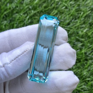 May include: A rectangular, emerald-cut gemstone with a vibrant, translucent light blue colour. The stone features sharp, clean facets that reflect light, creating a clear, glassy appearance. It is held against a background of green grass.
