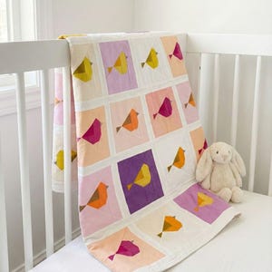 Könnte beinhalten: Ein bunter Patchwork-Quilt mit geometrischen Vogelmustern in Gelb-, Orange-, Rosa- und Lilatönen. Der Quilt ist über ein weißes Kinderbett drapiert, mit einem Stoffhasen-Spielzeug in der Nähe. Der Quilt besteht aus Quadraten mit Vogelmotiven.