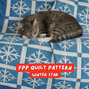 May include: A sleeping gray tabby cat rests on a blue and white quilt with a snowflake pattern. The quilt has a red banner that reads "FPP QUILT PATTERN WINTER STAR". The cat is curled up, with its eyes closed.
