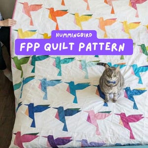 May include: A white quilt featuring a repeating design of origami-style hummingbirds in various colours, including orange, yellow, blue, pink, and purple. The text "HUMMINGBIRD FPP QUILT PATTERN" is on a purple banner. A grey tabby cat is on the quilt.