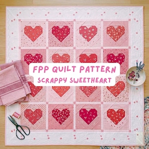 Op de afbeelding: Een handgemaakte quilt met een hartpatroon in roze en rode tinten. De quilt heeft de tekst "FPP QUILT PATTERN SCRAPPY SWEETHEART". De quilt is omgeven door een witte rand met een kanten ontwerp. Een schaar, draad en een kleine kom met bloemen zijn ook zichtbaar.