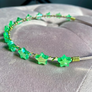 May include: A headband featuring a series of translucent, bright green star-shaped beads. The stars are attached to a thin, metallic band using a coiled, lime green wire that wraps around the frame between each individual star element.