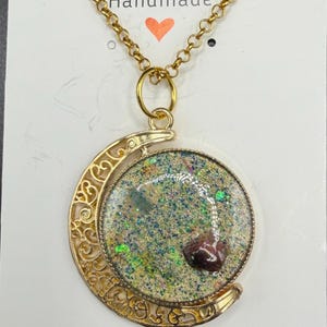 May include: Gold-toned necklace with a pendant featuring a glitter-filled circular design and a crescent moon frame. The pendant has a dark red stone and the chain is delicate. The word "Handmade" is printed at the top.