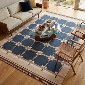 Op de afbeelding: Een rechthoekig vloerkleed met een geometrisch patroon in marineblauw, beige en bruin. Het vloerkleed is geplaatst in een woonkamer met een bank, salontafel en stoelen. Het ontwerp van het vloerkleed heeft een rasterachtig patroon.